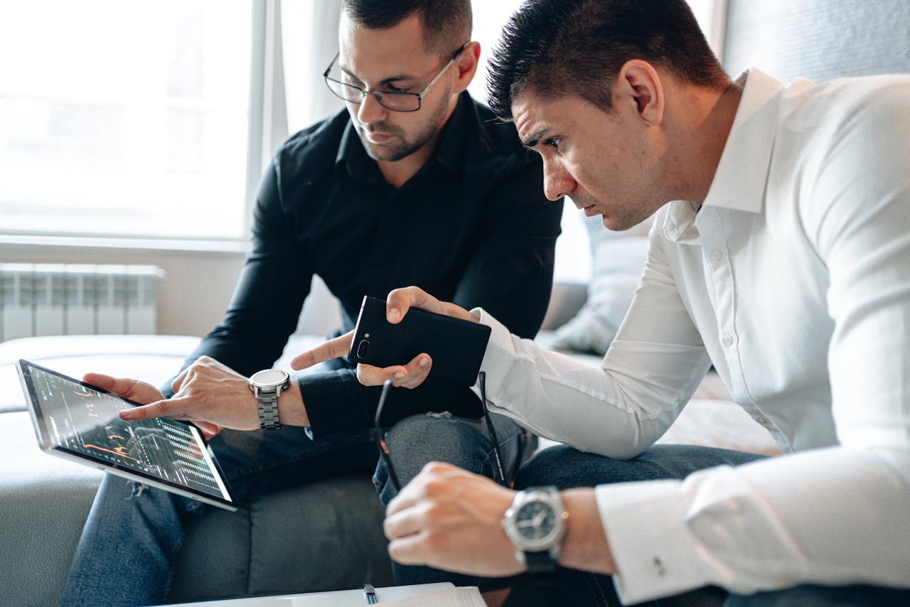 services-02 Two men analyzing financial data on a digital tablet indoors in a professional setting.
